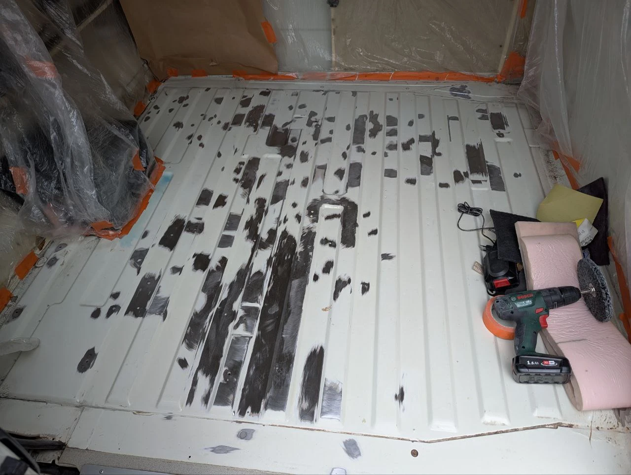 The image shows the cargo area floor of an old VW bus. The white paint has been taken off in many places. Around it, the walls are taped off with foil and masking tape. A cordless drill with a large sanding disc lies to the right.