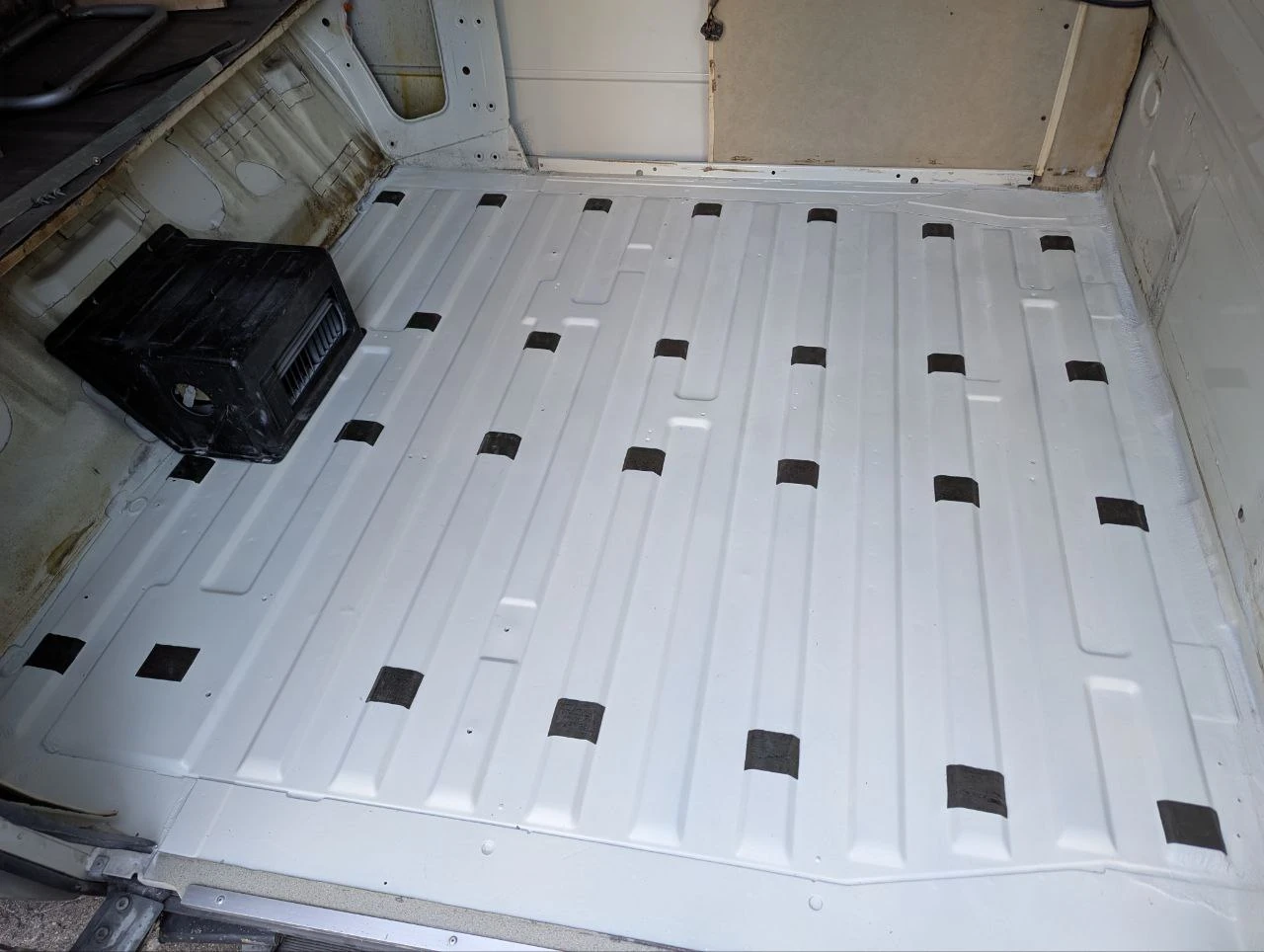The image again shows the cargo area floor of the VW bus. The paint is now renewed and uniformly white. Short black strips of tape are glued distributed over the raised seams of the floor.